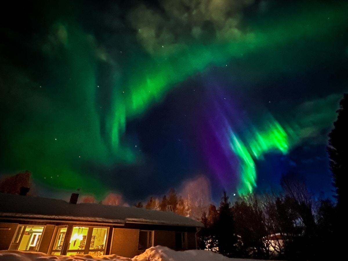 The night sky is illuminated by vibrant auroras, showcasing swirling patterns of green and purple hues. A modest house is visible in the foreground, with warm light spilling from its windows, creating a cozy contrast against the natural display above.