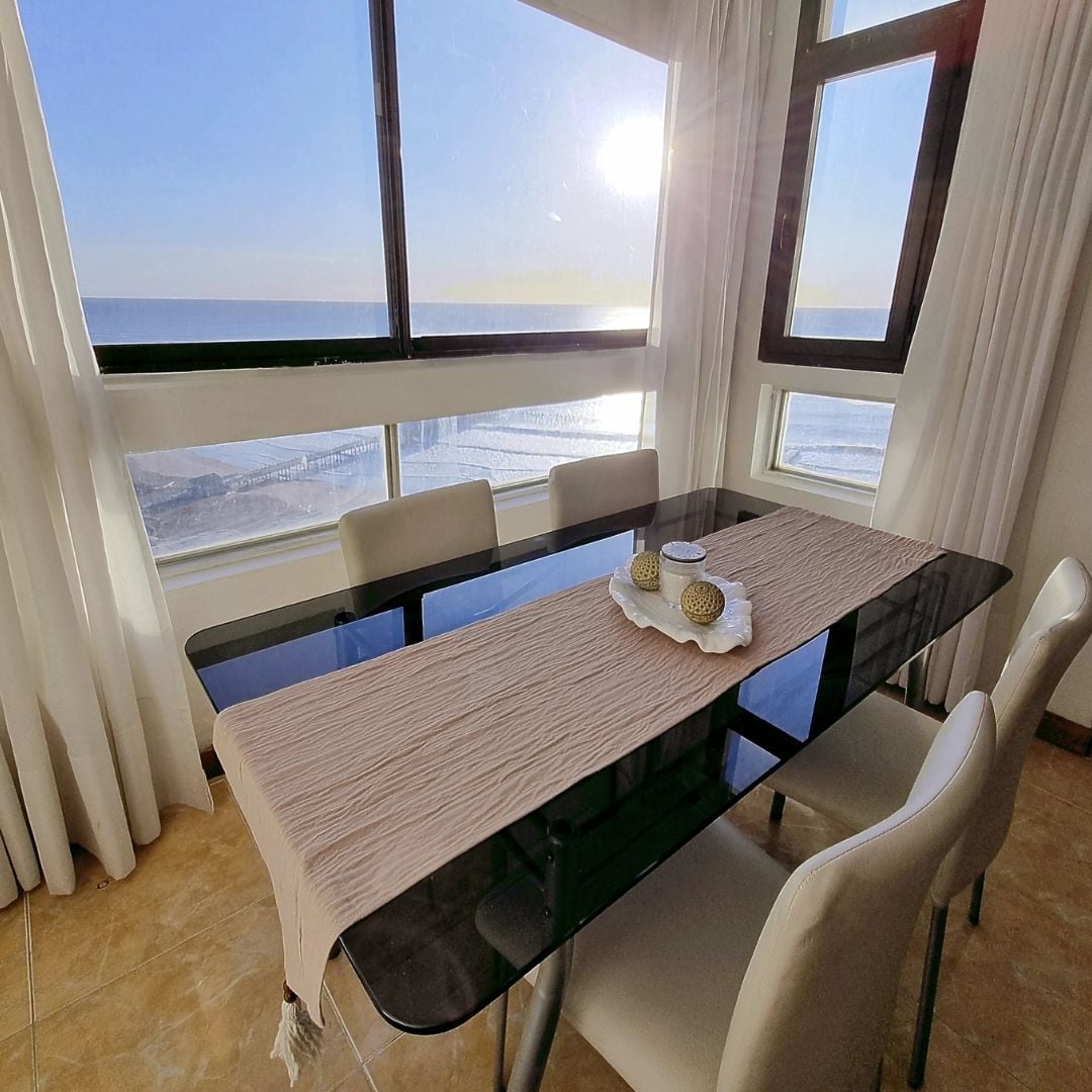 A dining space is illuminated by sunlight streaming through large windows, offering a view of the ocean. A dark glass table sits centered, adorned with a neutral table runner and decorative items. Four light-colored chairs surround the table, creating an inviting space for meals.