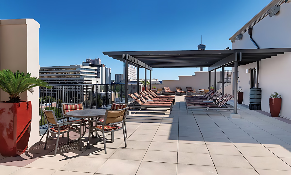 A rooftop terrace features a shaded seating area with a round table and chairs, surrounded by lounge chairs arranged along the edge, offering views of the city skyline. Potted plants add greenery to the space, enhancing the outdoor atmosphere.