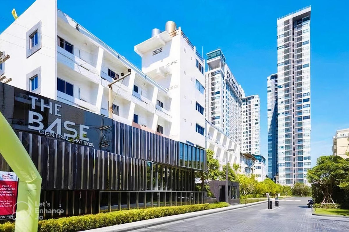 The exterior of The Base Central Pattaya is presented, featuring a modern building with large glass facades. The entrance is marked with the property name, and neighboring buildings rise in the background against a clear blue sky. Lush greenery lines the pathway leading to the entrance.