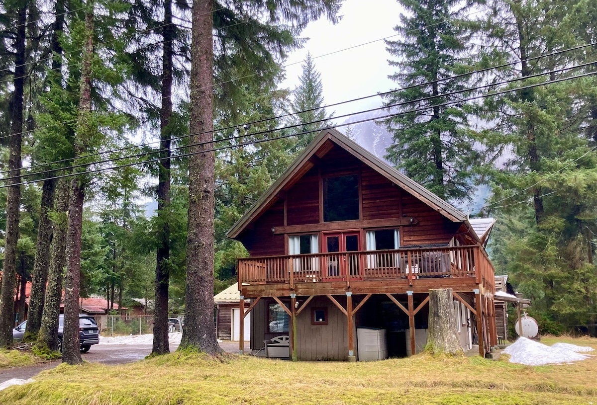 A cedar lodge style home is set among tall trees, featuring a wrap-around balcony that offers outdoor space. The structure displays a gabled roof and large windows, allowing natural light to enter the interior. Snow remains visible on the ground, indicating a seasonal charm.