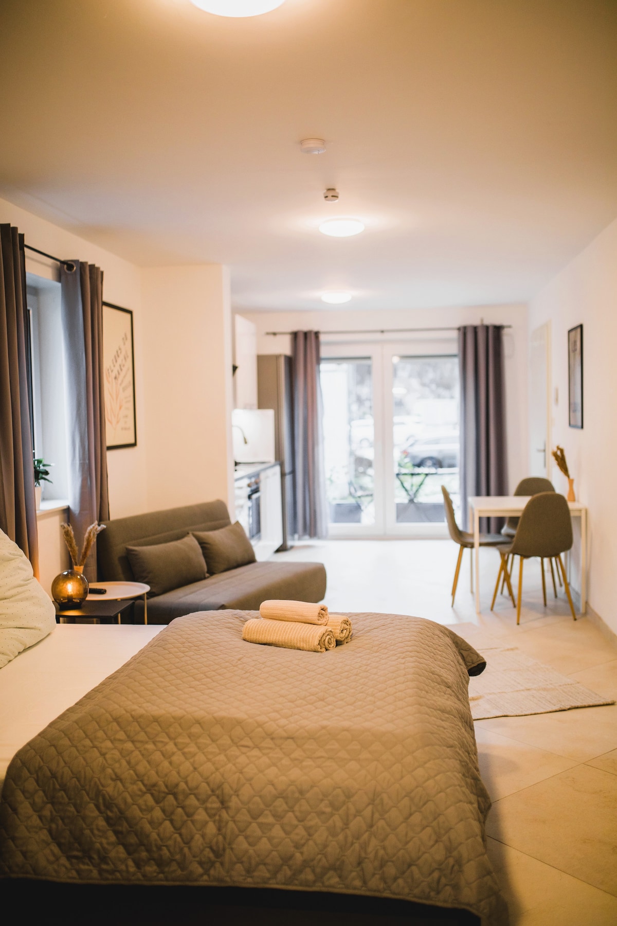 A studio apartment is depicted, featuring a spacious bed with a quilted cover. An inviting sofa is positioned beside a dining table with two chairs. Natural light enters through large glass doors, leading to a small outdoor terrace. Neutral-colored walls enhance the open atmosphere.