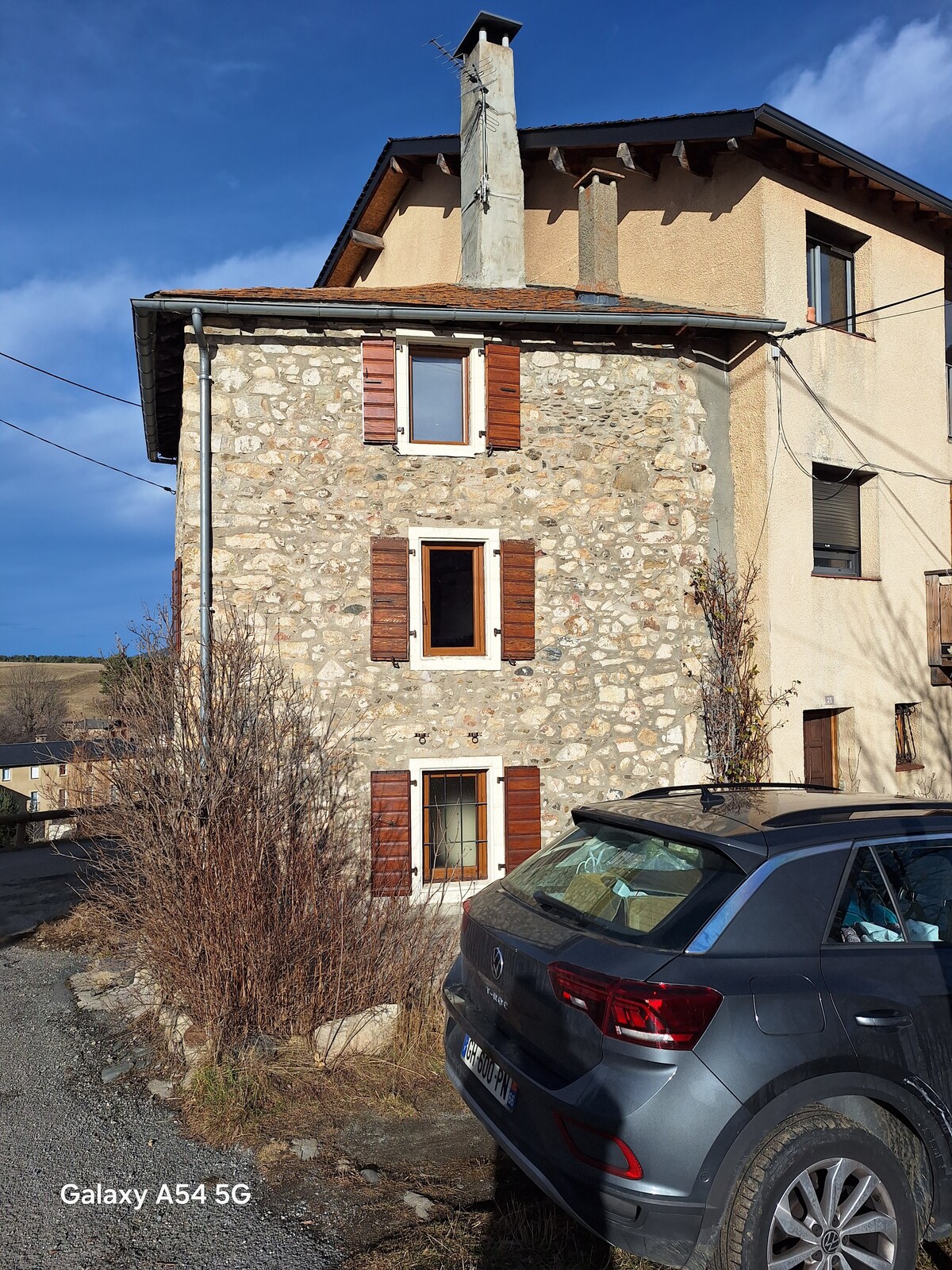 The exterior of a multi-level stone house is showcased, featuring wooden window shutters and a chimney. A car is parked nearby, and the surrounding landscape includes some shrubbery, suggesting a rural setting. Clear blue skies create a bright backdrop.