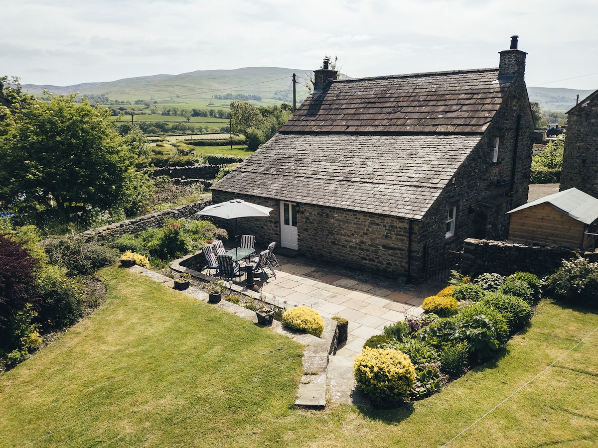 A stone cottage is set in a lush green landscape, featuring a well-maintained garden with blooming yellow flowers. A stone patio offers outdoor seating with chairs and an umbrella, inviting views of the rolling hills in the background under a clear sky.