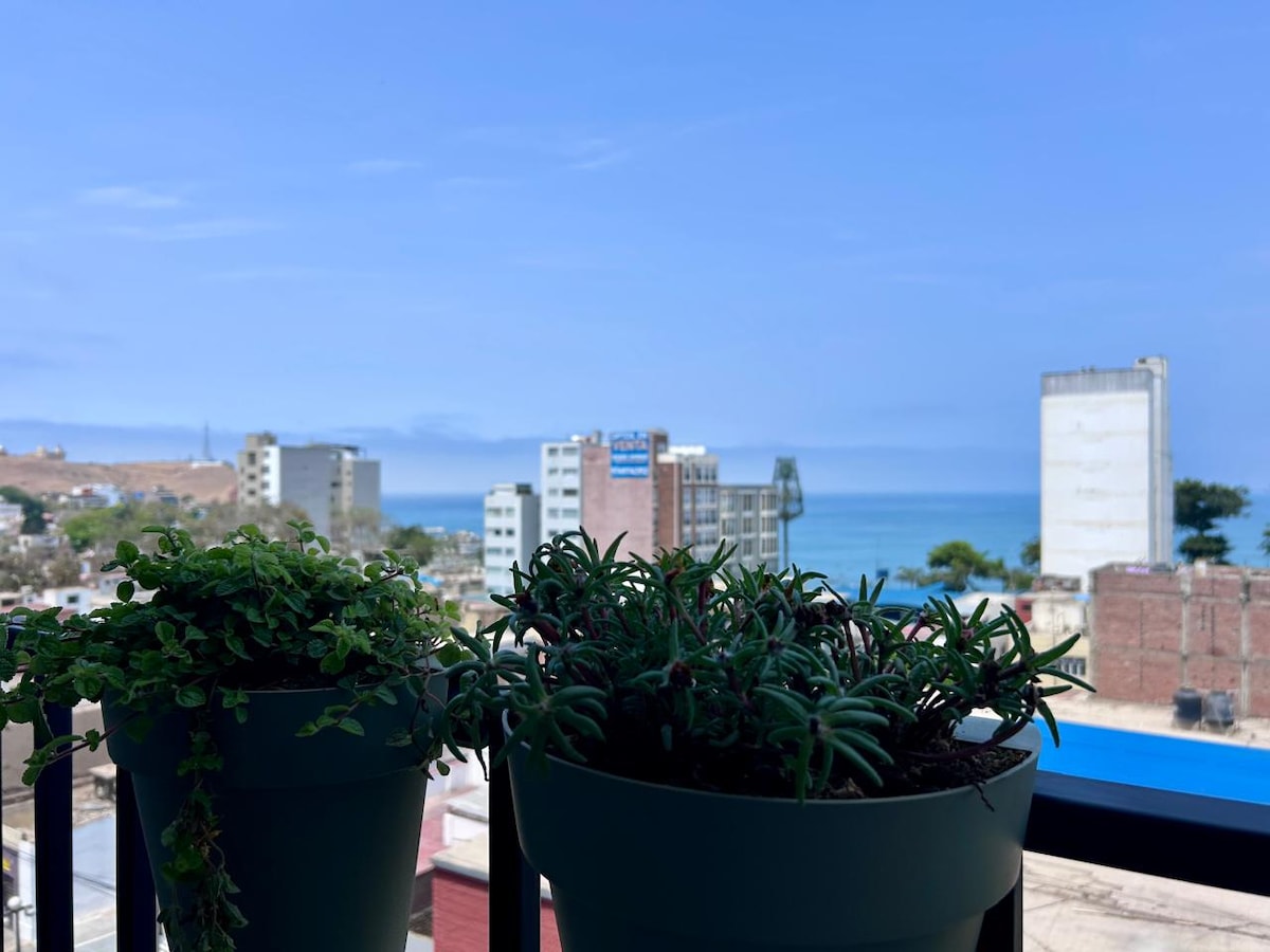 A view from the balcony showcases vibrant green potted plants in the foreground, while the horizon reveals a blend of urban buildings meeting the serene blue ocean under a clear sky. The tranquility of the landscape is enhanced by the inviting atmosphere.