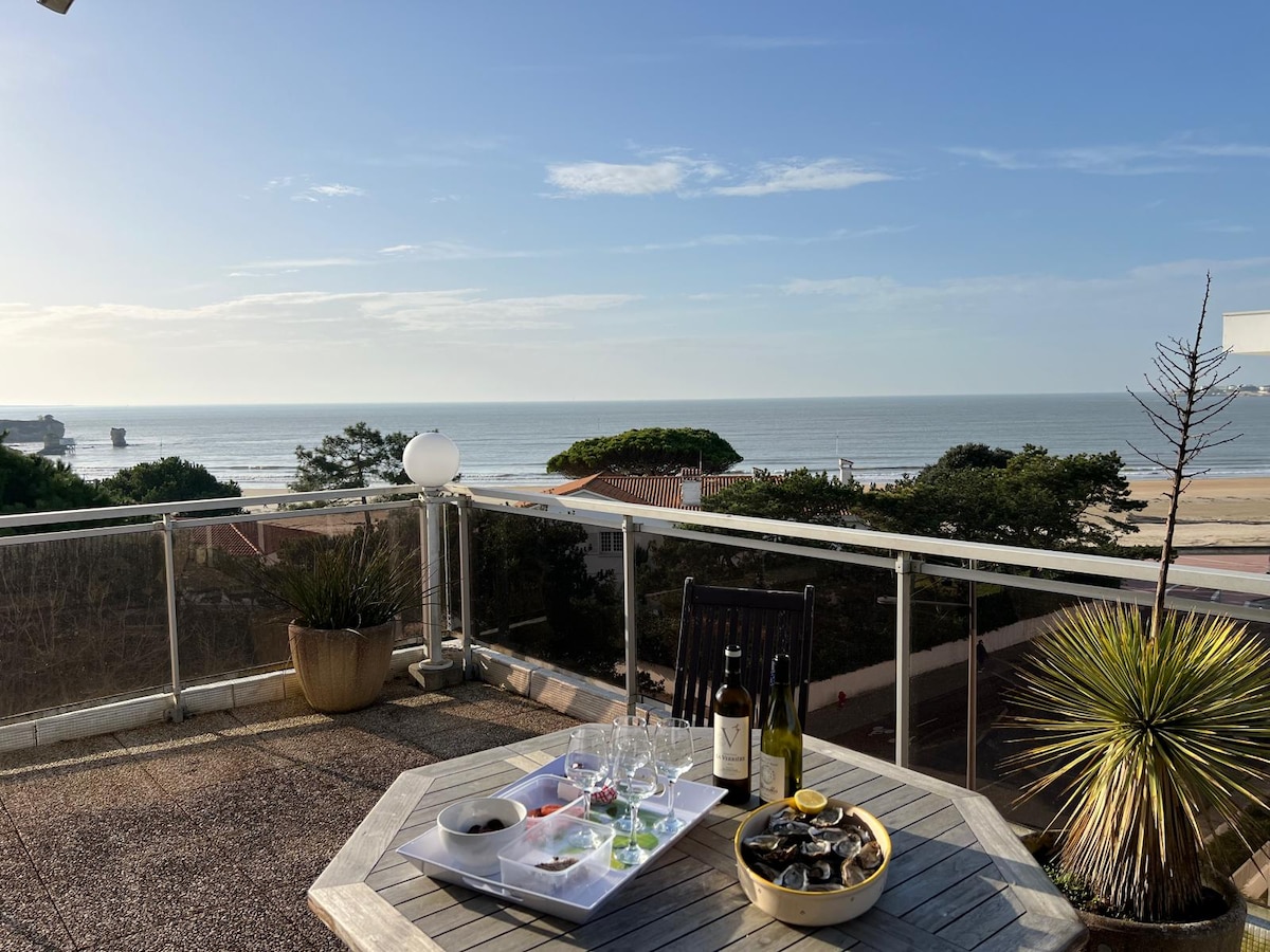 An outdoor dining table is set for an ocean view meal, featuring glasses and bottles of wine, along with a platter of oysters. The tranquil sea and coastal trees are visible in the distance under a clear sky, highlighting the spacious terrace area.