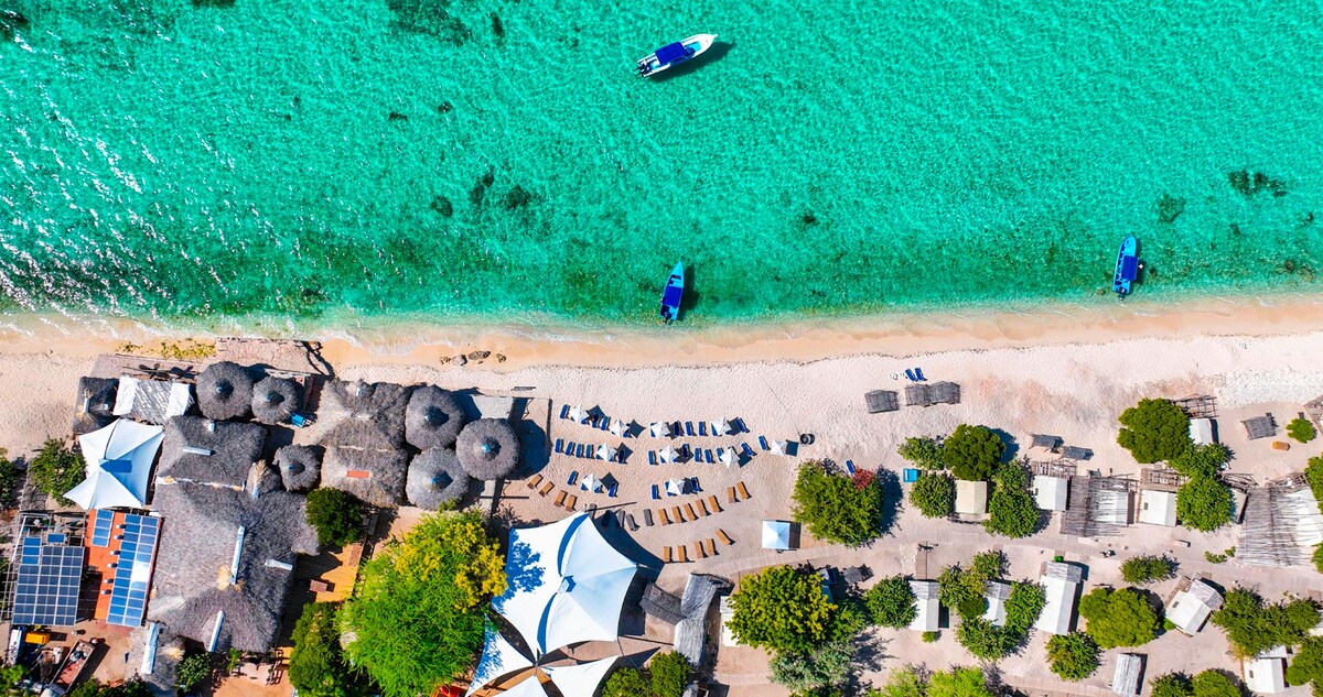 An aerial view exposes a vibrant beach scene with turquoise waters lapping at the shore. Sun loungers are arranged along the sandy beach, while small boats are anchored nearby. Beach umbrellas are positioned among lush greenery and shaded areas, contributing to the lively coastal atmosphere.