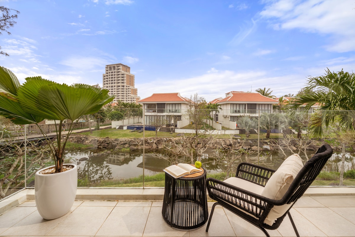 A cozy outdoor seating area is depicted, featuring a black wicker chair and a small side table with a book and a drink. The view reveals lush greenery and a calm river, framed by modern villas in the background under a clear blue sky.