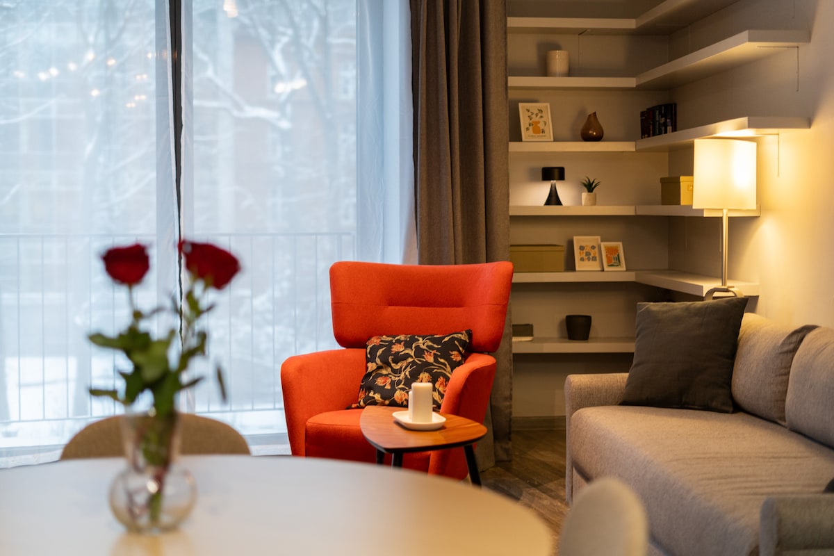 A vibrant orange armchair is positioned beside a small side table, featuring a candle. A soft gray sofa complements the decor, while a round dining table with a vase of roses is visible. Shelves display decorative items against a light gray wall backdrop.