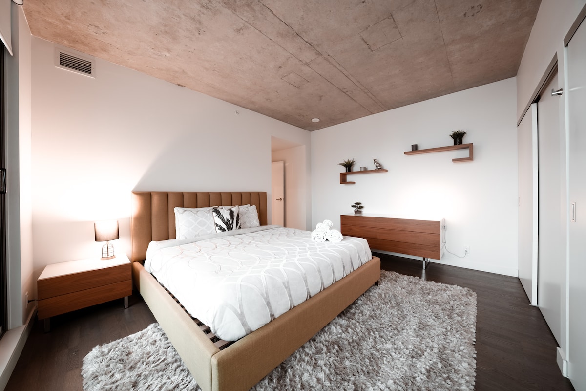 A modern bedroom features a spacious bed with a light-colored coverlet, situated on a soft rug. Nightstands on either side of the bed are topped with lamps. A wooden shelf adorns the wall, holding decorative plants, while sliding doors provide storage options.