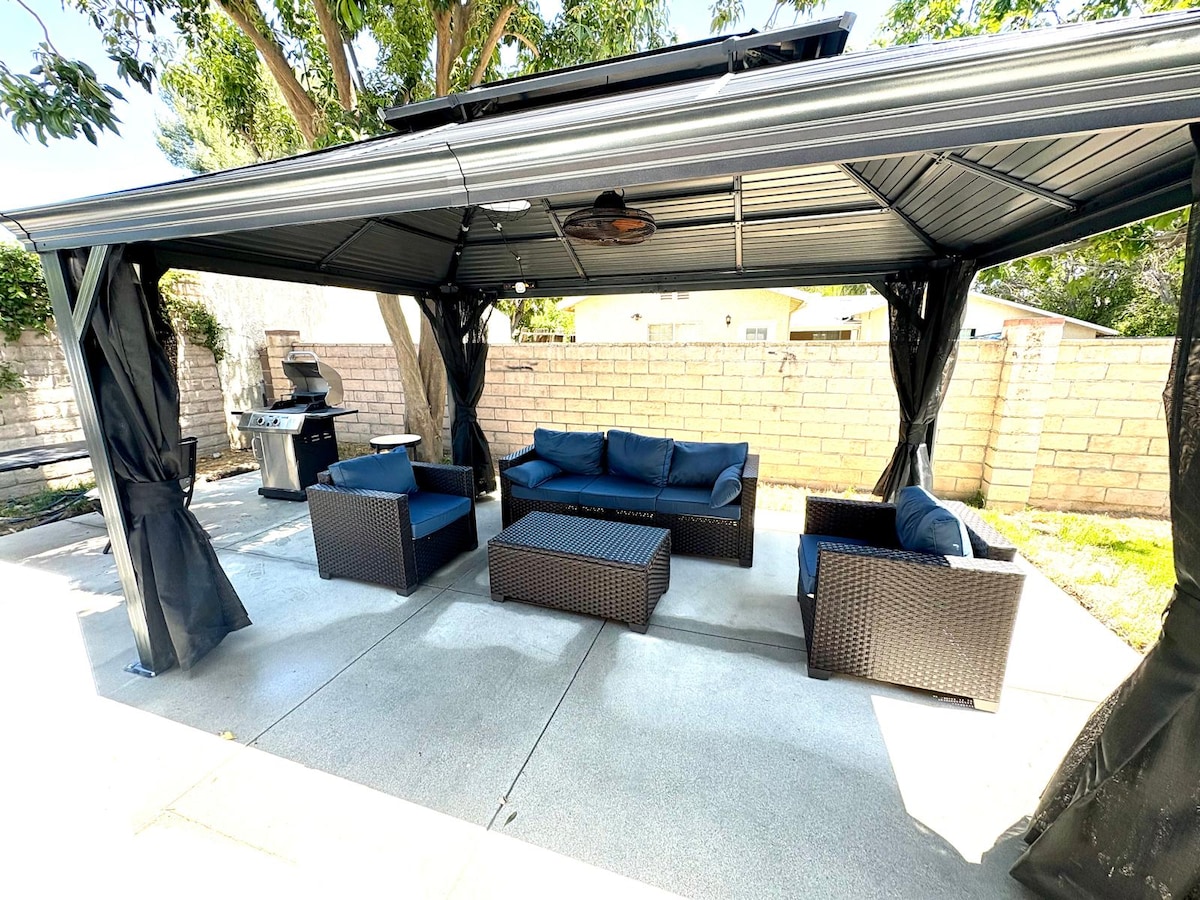 An outdoor gazebo is featured, providing a shaded seating area with a sectional sofa and matching chairs, all in dark wicker. A coffee table sits in the center, and a barbecue grill is positioned to the left against a stone wall, surrounded by a neatly landscaped yard.