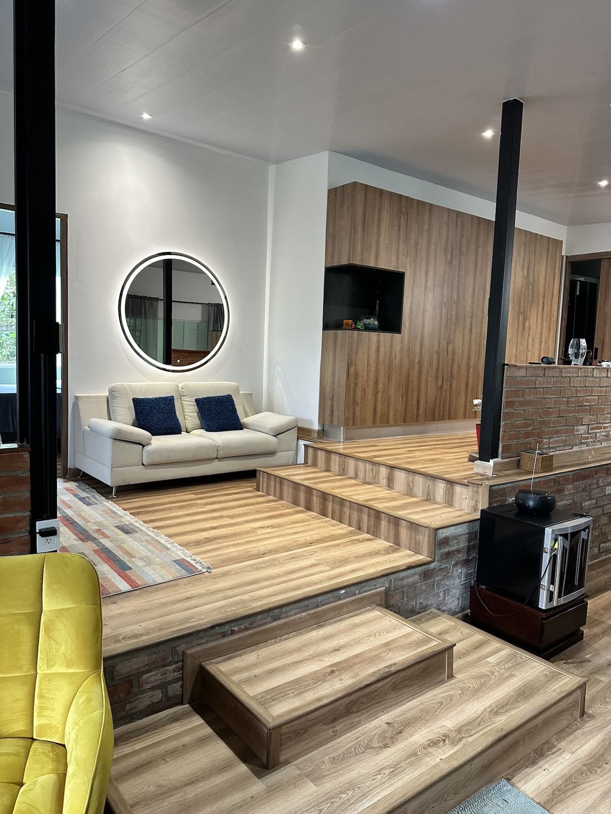 A living area is showcased with a light-colored sofa placed near a round mirror on the wall. Steps lead down to a warm wooden floor, while a brick wall provides texture. A cozy yellow chair complements the space, accompanied by a media unit in the background.