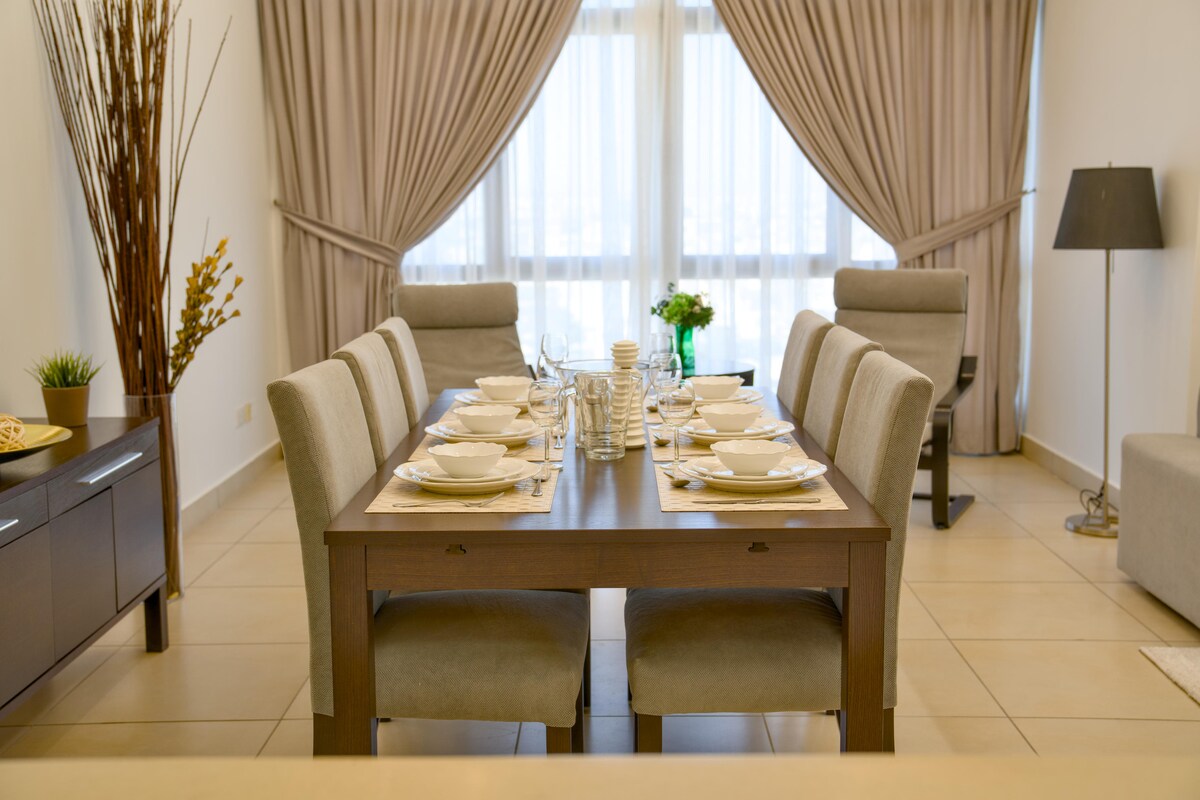 A dining area is featured with a long wooden table set for eight. Light-colored chairs surround the table, which is adorned with plates and glassware. Sheer curtains drape over the large windows, allowing natural light to filter into the space.