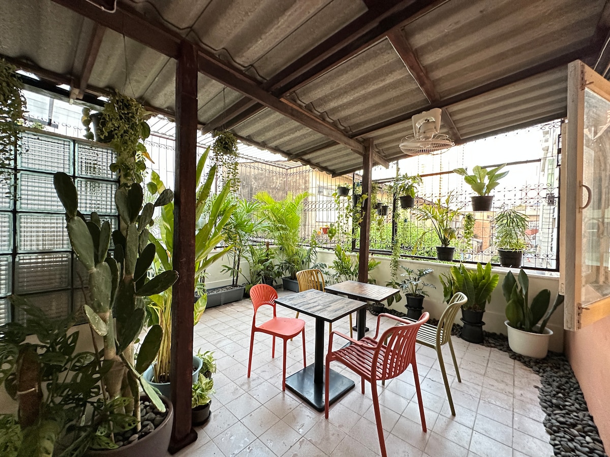 A spacious rooftop garden area is featured, surrounded by a variety of potted plants and greenery. Four brightly colored chairs are arranged around a dark wooden table. Natural light filters through the overhead structure, creating a peaceful atmosphere.