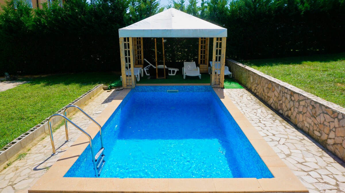 An inviting outdoor pool is surrounded by a well-maintained garden, featuring a shaded seating area with sun loungers. A sturdy pool ladder is positioned at one end, and the tranquil blue water glistens under natural light.