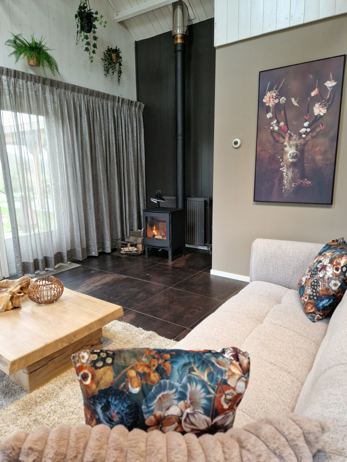 A welcoming living area features a light-colored sofa adorned with patterned cushions. A wooden coffee table is positioned centrally, complementing the cozy atmosphere. A black wood stove stands against a dark wall, with a nature-themed artwork displayed nearby. Soft curtains frame expansive glass doors leading to the outside.