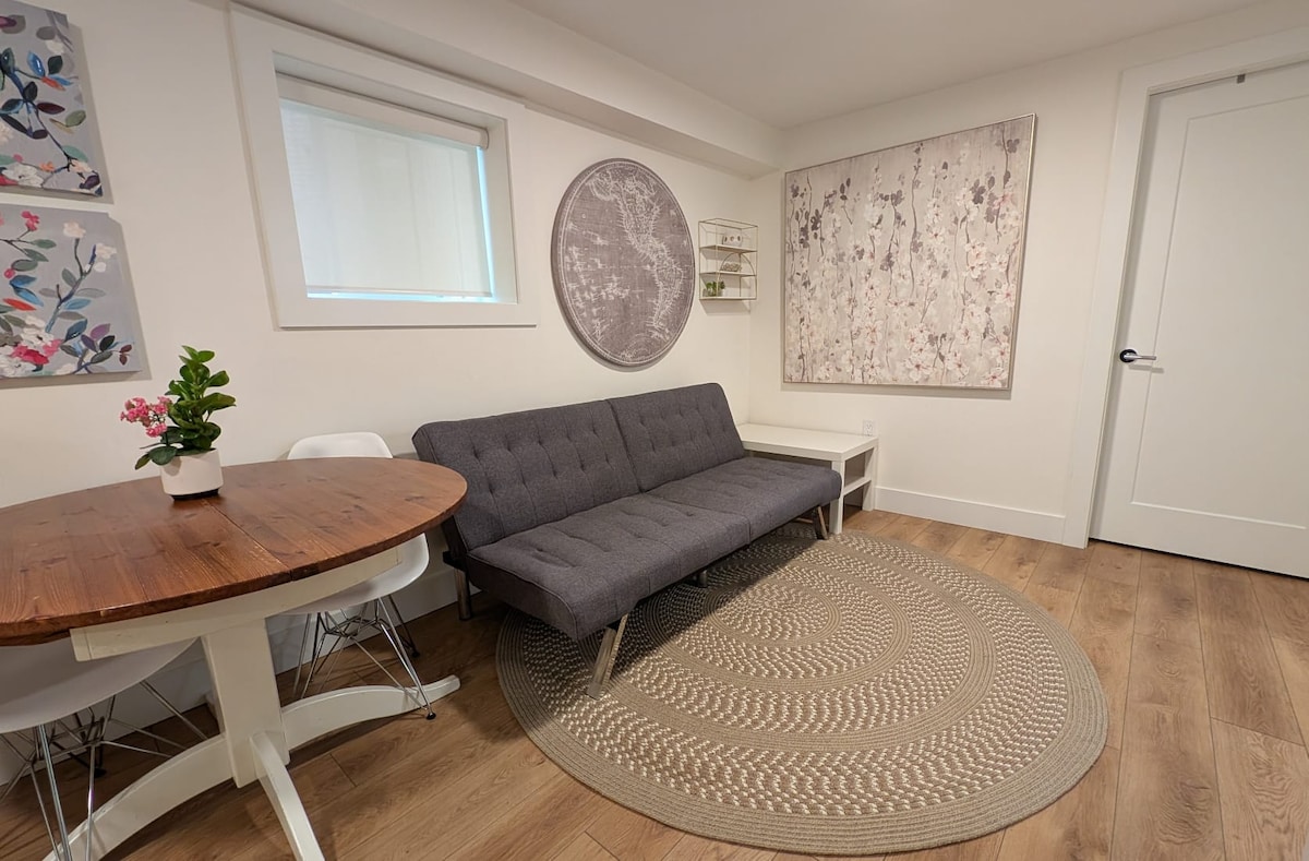 A modern living area features a grey futon couch positioned beside a round table with white chairs. A woven circular rug enhances the space, while two decorative wall hangings add a touch of style. Natural light filters in through a nearby window, creating a bright ambiance.