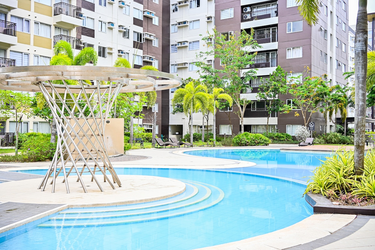 The image shows a serene pool area surrounded by well-maintained gardens and palm trees. A modern structure is positioned over the shallow end, while lounge chairs are seen placed along the perimeter of the pool, inviting relaxation in a tranquil environment.