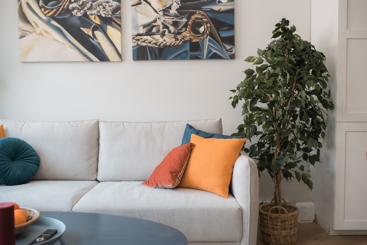 A modern living space features a light-colored sofa adorned with vibrant pillows in orange and teal. Two pieces of artwork hang on the wall above, complemented by a decorative plant positioned in a woven basket, adding a touch of greenery to the room.