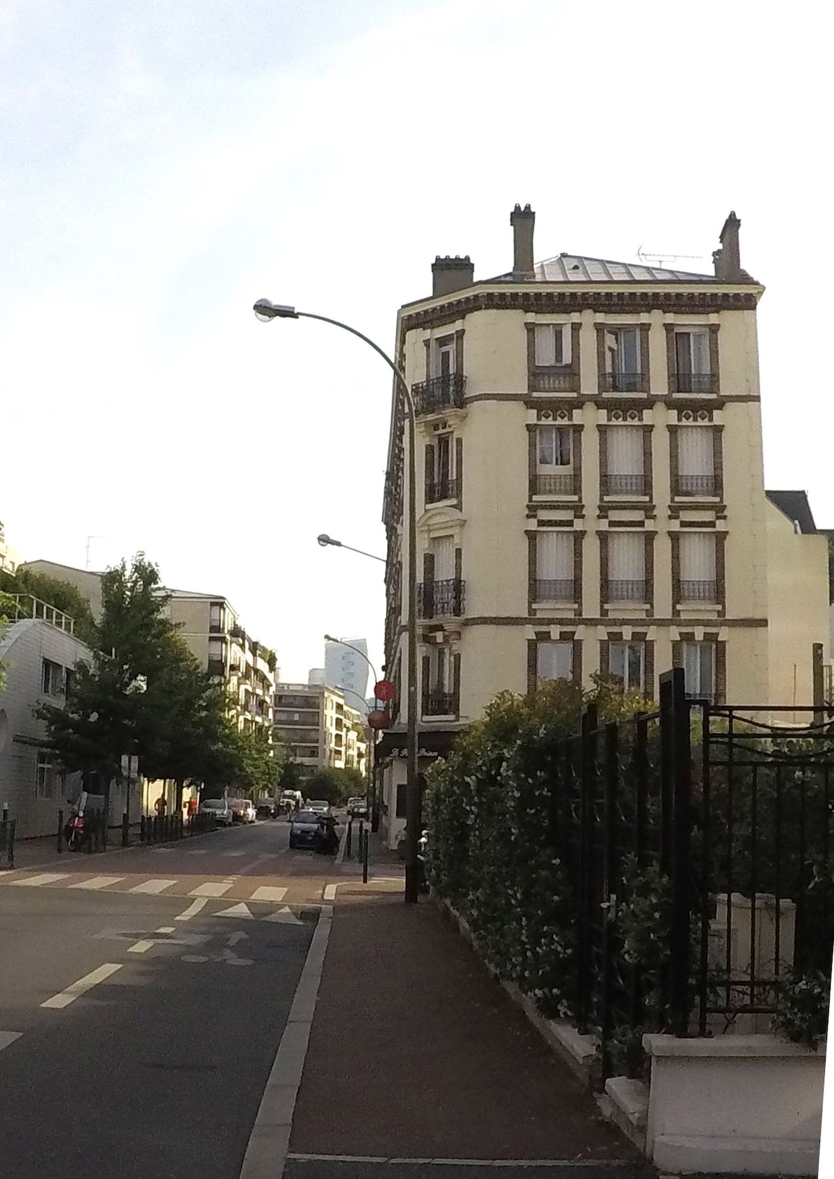A street view capturing a classic four-story building with a light-colored facade and multiple windows. The street is lined with trees and parking spaces, with a glimpse of modern architecture in the background. Sidewalks are present for pedestrians, enhancing the urban setting.