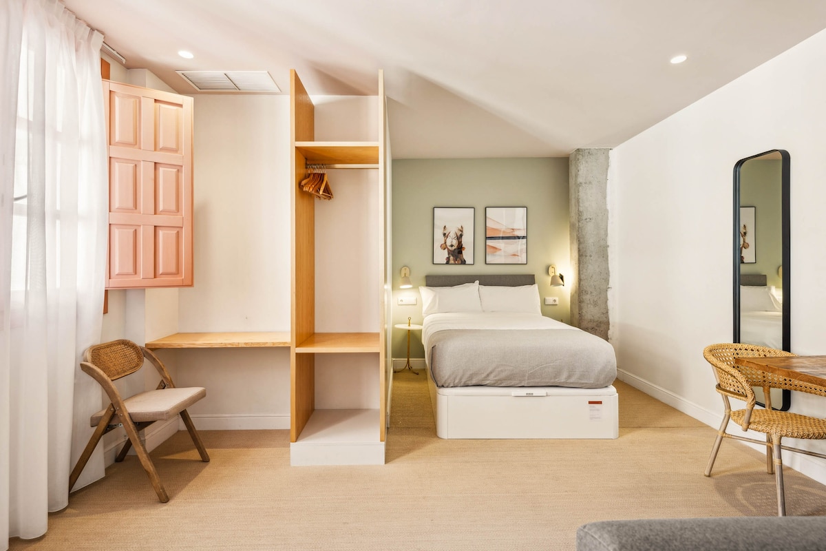 A spacious studio layout features a comfortable bed with soft bedding and a light grey duvet. A minimalist open wardrobe with a pink door stands nearby, while a small writing desk and a chair are positioned in one corner, enhancing the functional space.