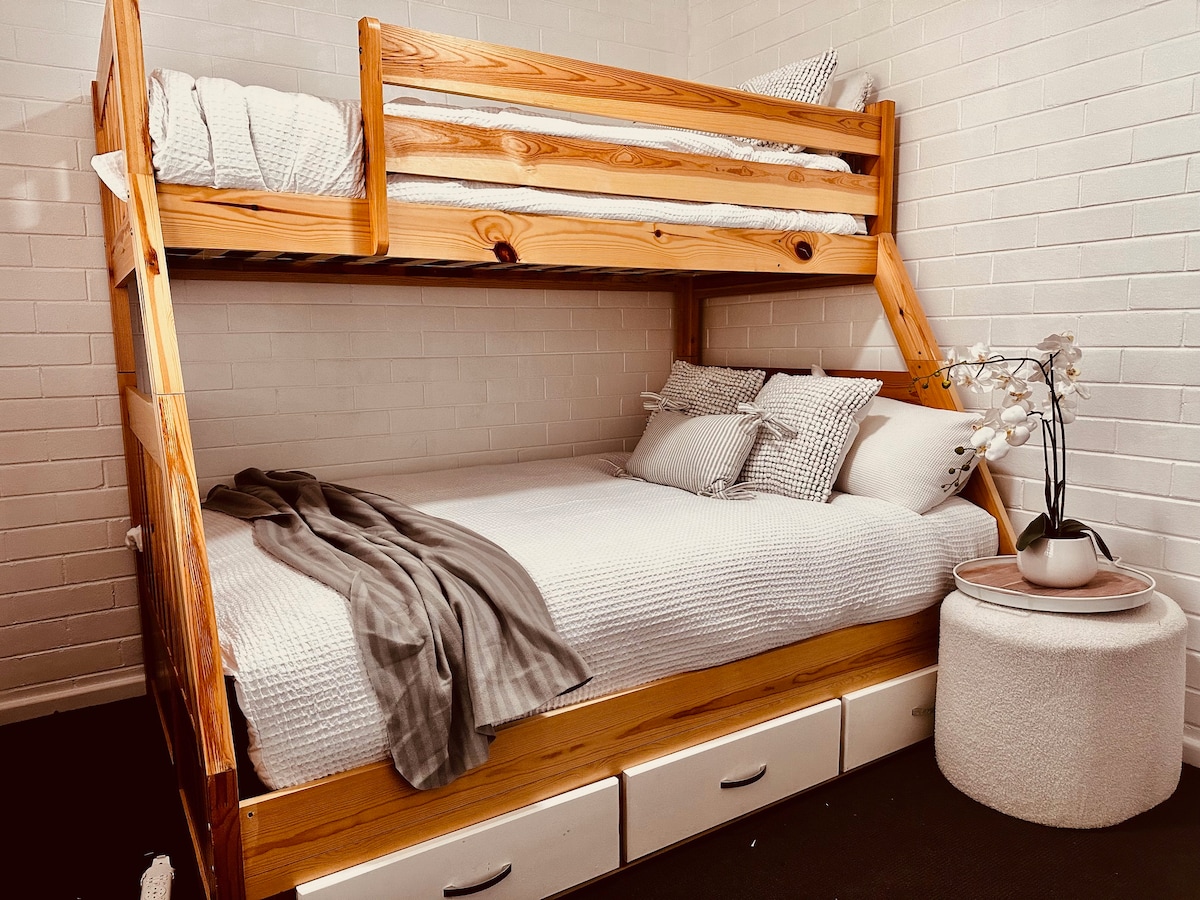 A compact bedroom features a wooden bunk bed with a spacious lower bed and a top bunk. Soft, white bedding is layered with decorative pillows. A cozy throw lies across the lower bed, and a round side table holds a small vase with white flowers.