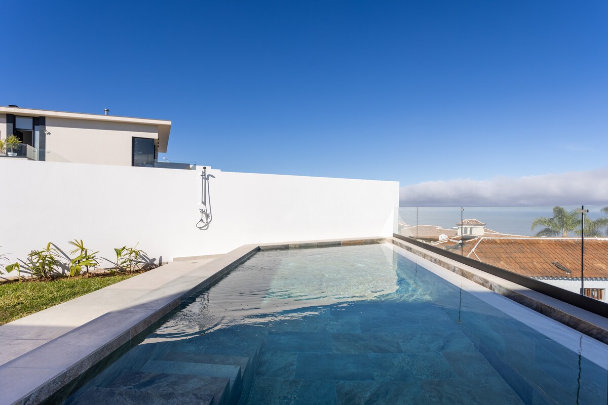 An infinity pool with clear water is positioned against a white wall, offering unobstructed views of the sky. Lush greenery is visible on the pool's edge, contributing to the serene atmosphere. The surrounding area showcases a clean, modern design.