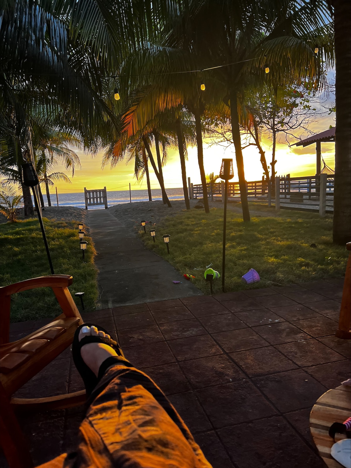 A serene seating area is shown with wooden rocking chairs facing a vibrant sunset over the ocean. Palm trees are visible framing the scene, while the path leading to the beach is lined with soft lighting. The tranquil atmosphere is highlighted by a grassy area nearby.