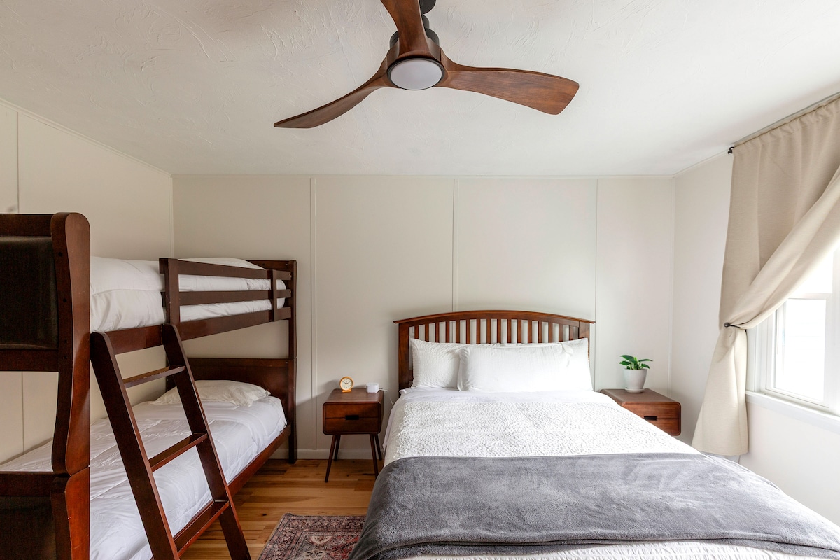 A room features a spacious Queen bed and twin bunk beds, all adorned with soft bedding. Natural light enters through a window, complemented by a light curtain. A small nightstand is situated beside the bed, and a potted plant adds a touch of greenery.