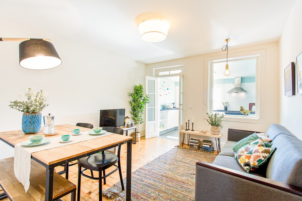 A welcoming living area features a dining table set for four, a cozy sofa adorned with decorative cushions, and a flat-screen TV. Natural light enters through large windows, illuminating the space. A well-equipped kitchen is visible in the background, enhancing the homey atmosphere.