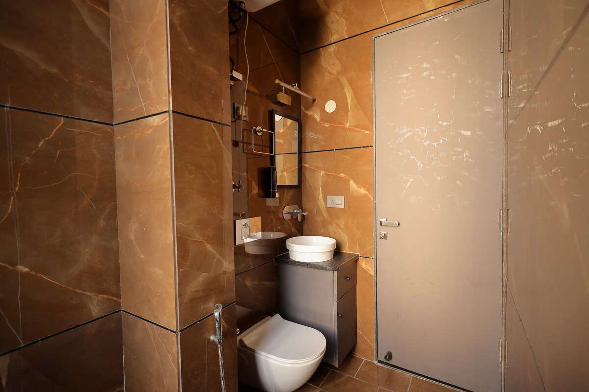 A modern bathroom features elegant brown marble walls with subtle veining. A round basin is positioned above a sleek cabinet, while a toilet is placed against the wall. A large mirror reflects the well-lit space. The door is a muted grey, offering a clean and simple design.