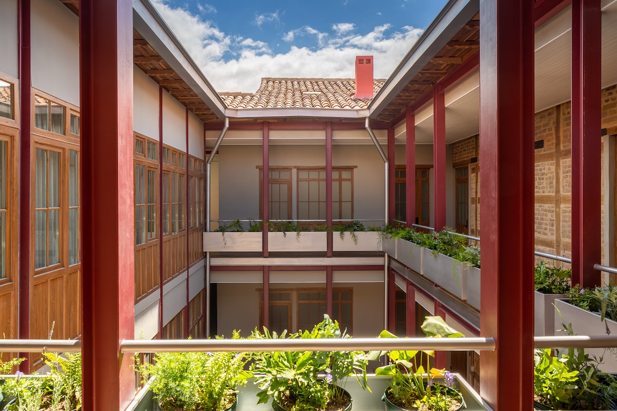 An open courtyard is surrounded by red pillars and features multiple levels of balconies with lush green plants. Large windows are visible in the surrounding walls, allowing natural light to flow through the space, reflecting the blend of modern design and historical architecture.
