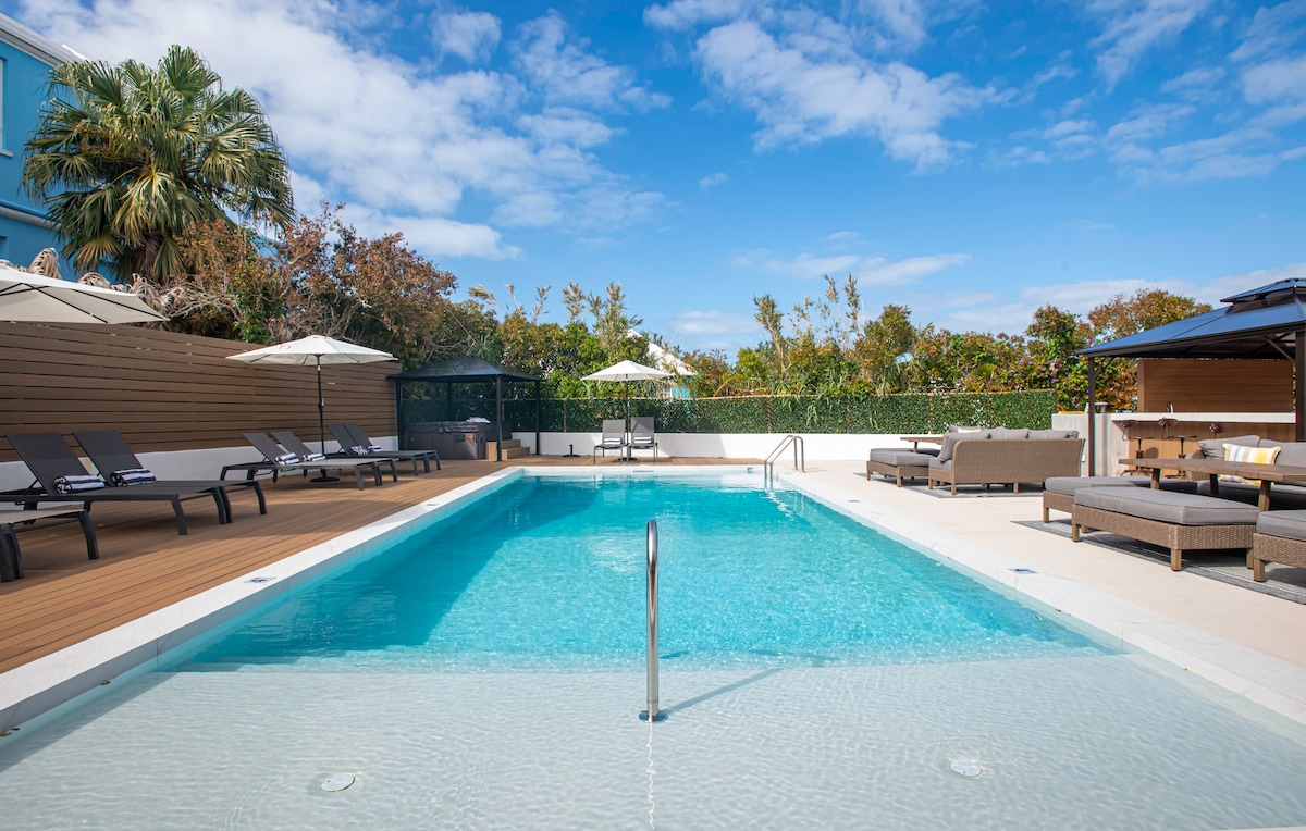 An outdoor pool area showcases a refreshing heated saltwater pool surrounded by lounge chairs and umbrellas. Lush greenery and palm trees line the perimeter, enhancing the tranquil setting under a bright blue sky.