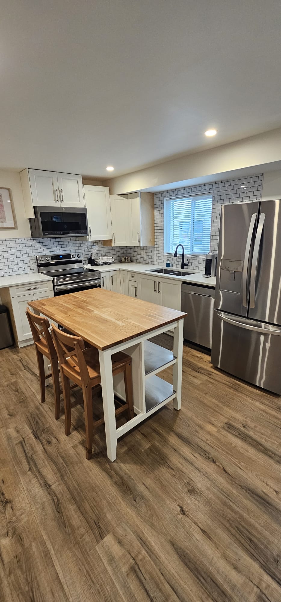 A modern kitchen features stainless steel appliances, including a refrigerator and gas stove. Light wood flooring complements the white cabinetry and a wooden island with seating for two. Natural light filters through the window above the sink, enhancing the bright and functional workspace.