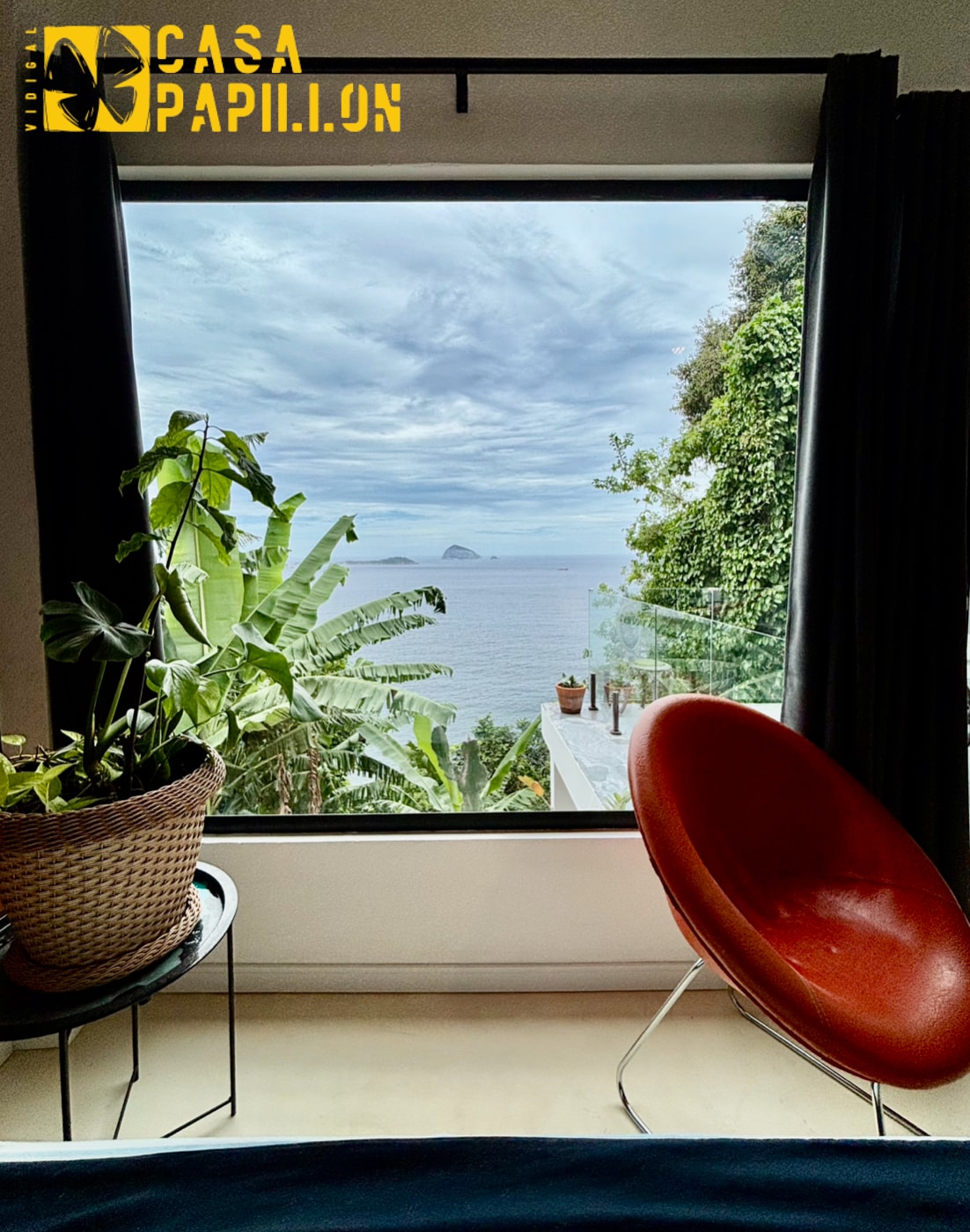 A comfortable interior showcases a large window offering views of the ocean and surrounding greenery. A potted plant sits on a side table next to a stylish chair, inviting relaxation while enjoying the natural landscape.