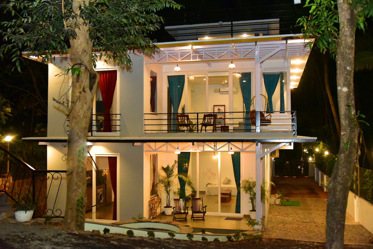 The two-story villa is illuminated at night, displaying a harmonious blend of indoor and outdoor spaces. Large glass doors open to a welcoming porch with comfortable seating. Colorful curtains frame the windows, enhancing the modern aesthetic of the exterior.