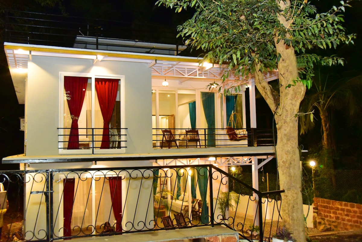 The two-level villa is showcased under night lighting, featuring a combination of large windows and terrace areas. Colorful drapes frame the windows, while outdoor seating is visible on both levels, promoting a relaxed gathering space amidst natural surroundings.