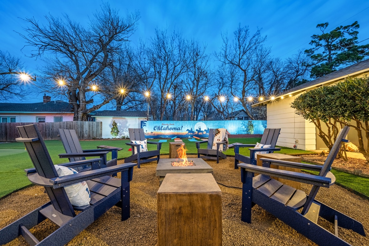 An outdoor space is illuminated by string lights, featuring comfortable seating around a fire pit. Four Adirondack chairs with cushions encircle a wooden table, creating a cozy gathering area. A mural depicting an Oklahoma landscape is visible in the background, adding a touch of character.