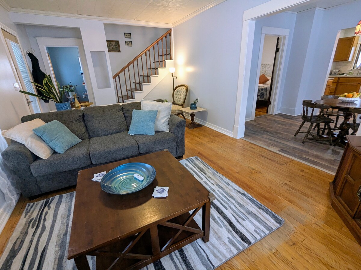 A cozy living area is shown with a charcoal gray sofa adorned with light blue pillows. A wooden coffee table with a decorative bowl is centered on a striped rug. The space is illuminated by natural light, showcasing a staircase and an adjoining dining area.