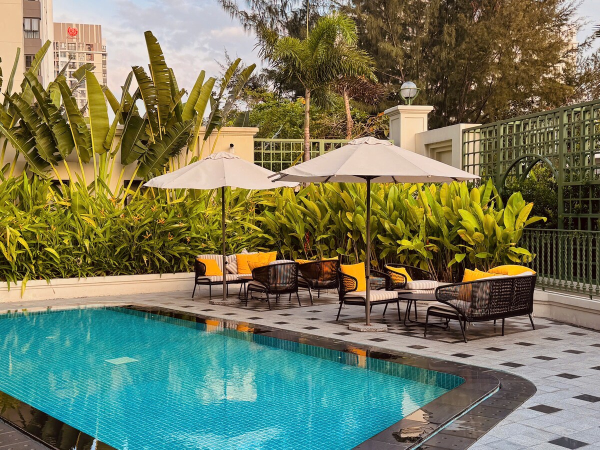 An outdoor area by the swimming pool is featured, surrounded by lush greenery. Comfortable seating arrangements with yellow cushions are positioned under large umbrellas. The pool's clear water reflects the sky, creating a serene atmosphere.