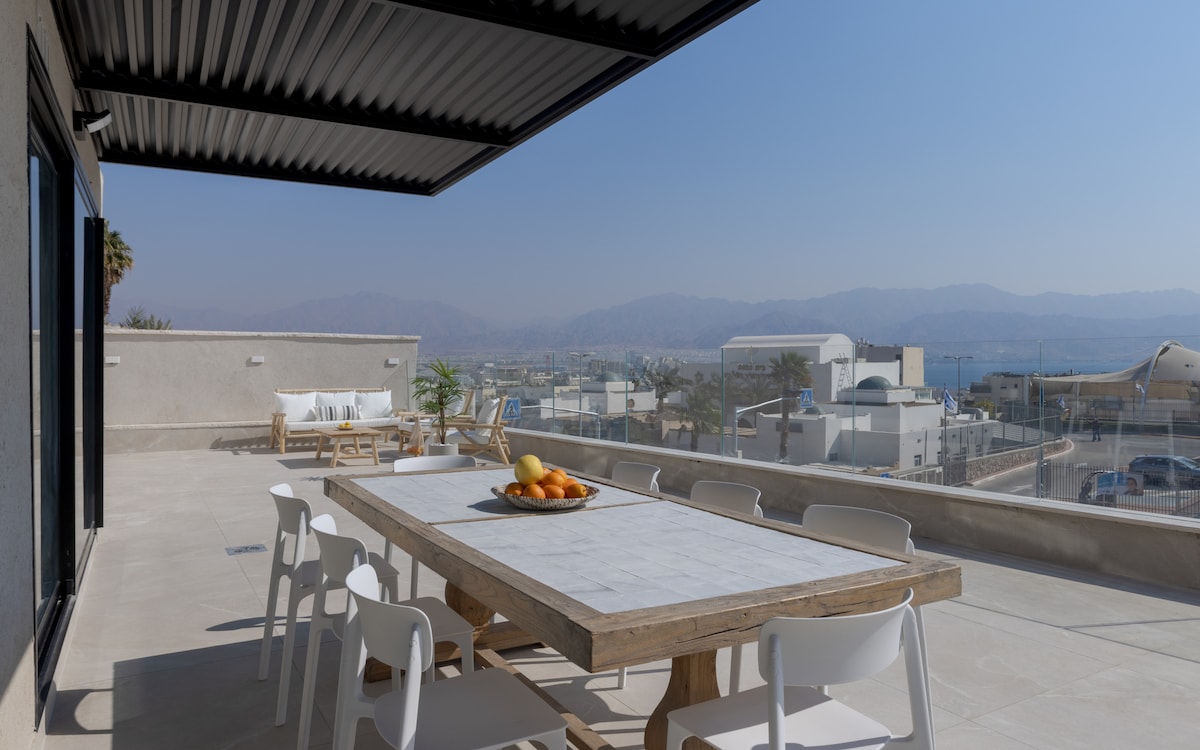 A spacious outdoor terrace features a large dining table surrounded by white chairs, offering a scenic view of the mountains and sea. A bowl of fresh fruit sits at the center of the table, ideal for enjoying meals al fresco.