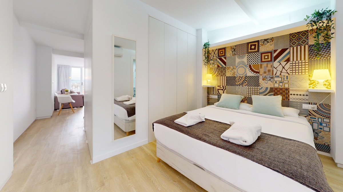 A spacious sleeping area features a bed with neutral linens and decorative pillows. A patterned accent wall adds visual interest. Soft lighting is provided by two lamps on each side of the bed, while a mirror reflects the room's inviting ambiance. The adjoining living space is visible in the background.