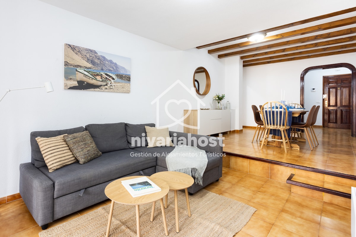 A spacious living area features a grey sectional sofa with decorative cushions, accompanied by a light wood coffee table. The space is well-lit, showcasing a dining area with a round table and wooden chairs in the background. Exposed wooden beams add warmth to the ceiling.