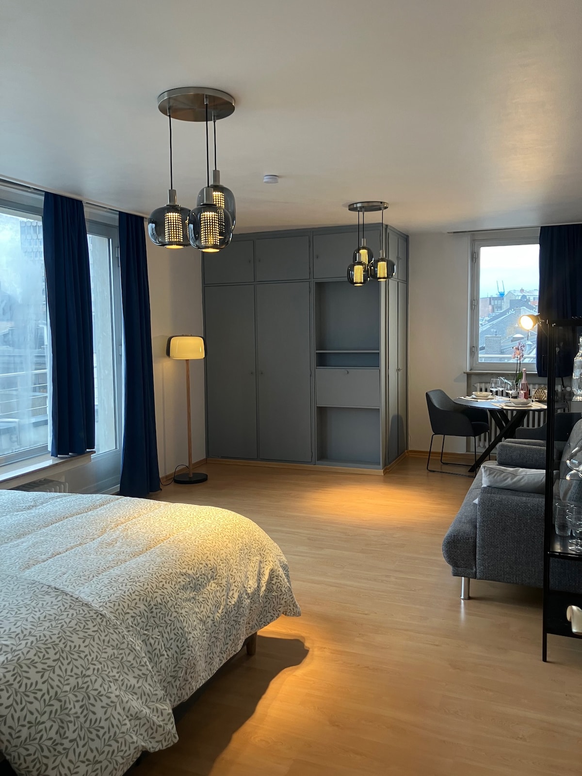 A well-appointed bedroom features a spacious bed with light-toned bedding. Soft lighting is provided by pendant fixtures and a floor lamp. A built-in wardrobe is visible, along with a dining area that includes a small table and modern chairs, under large windows with blue drapes.
