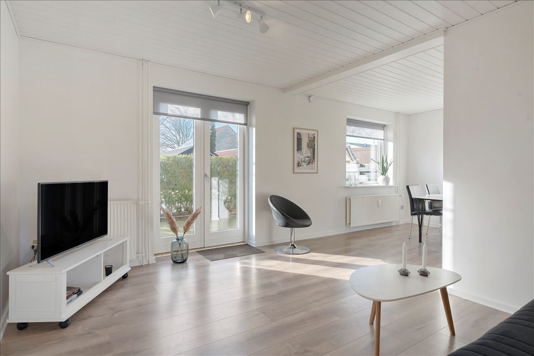 A bright living space features light-colored walls and wooden flooring. A television is positioned on a white cabinet, while a modern black chair and a round coffee table are present. Large sliding glass doors allow natural light to flood in, revealing a view of the garden.