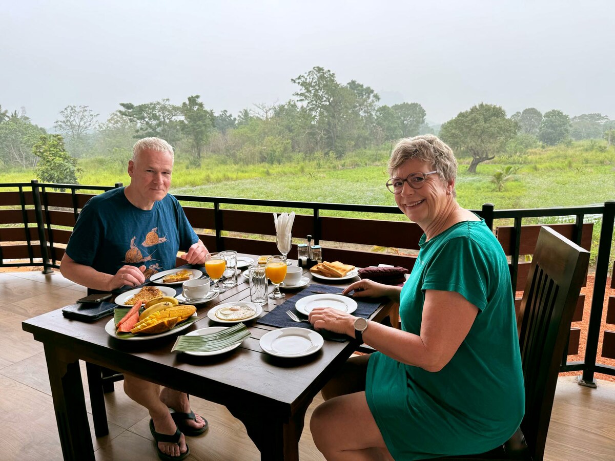 A dining table is set on a terrace overlooking a green landscape. Two plates of food, glasses of orange juice, and cutlery are arranged for breakfast. The scene includes a tranquil atmosphere, framed by wooden railings and a view of lush foliage in the background.