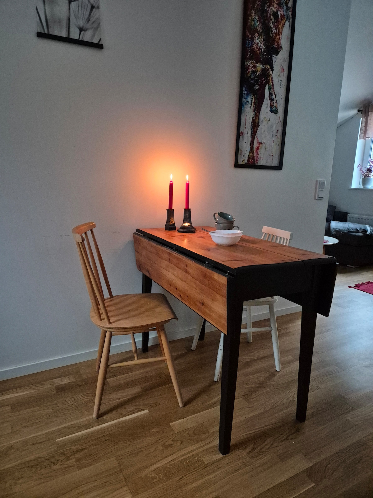 A wooden table with a natural finish is positioned against a wall. Two candles are lit, casting a soft glow. One light-colored chair and one wooden chair are placed beside the table. An abstract piece of wall art is visible in the background.