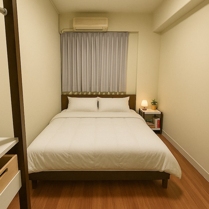 חדר השינה הזה משדר אווירה שלווה ומינימליסטית, עם מיטת קווין סייז במיקום מרכזי בגודל 60 אינץ' רוחב על 80 אינץ' אורך (152 ס"מ x 203 ס"מ). המיטה מעוטרת במצעים לבנים נקיים וכריות תואמות