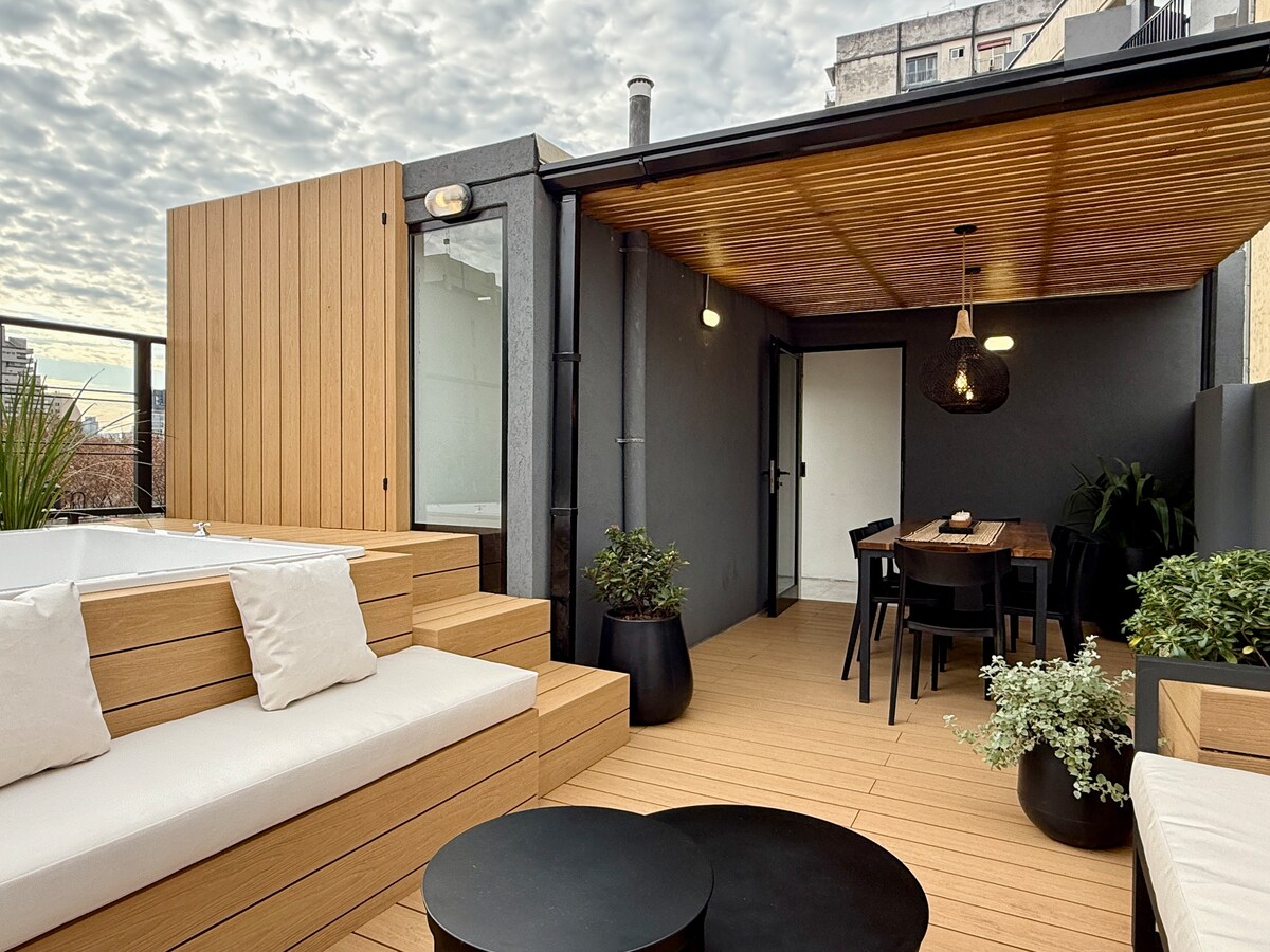 A private terrace features comfortable seating and a dining area under a covered wooden ceiling. Planters with greenery are placed around, enhancing the outdoor space. Soft cushions on the lounge seating invite relaxation, while the warm wood tones create a harmonious environment.