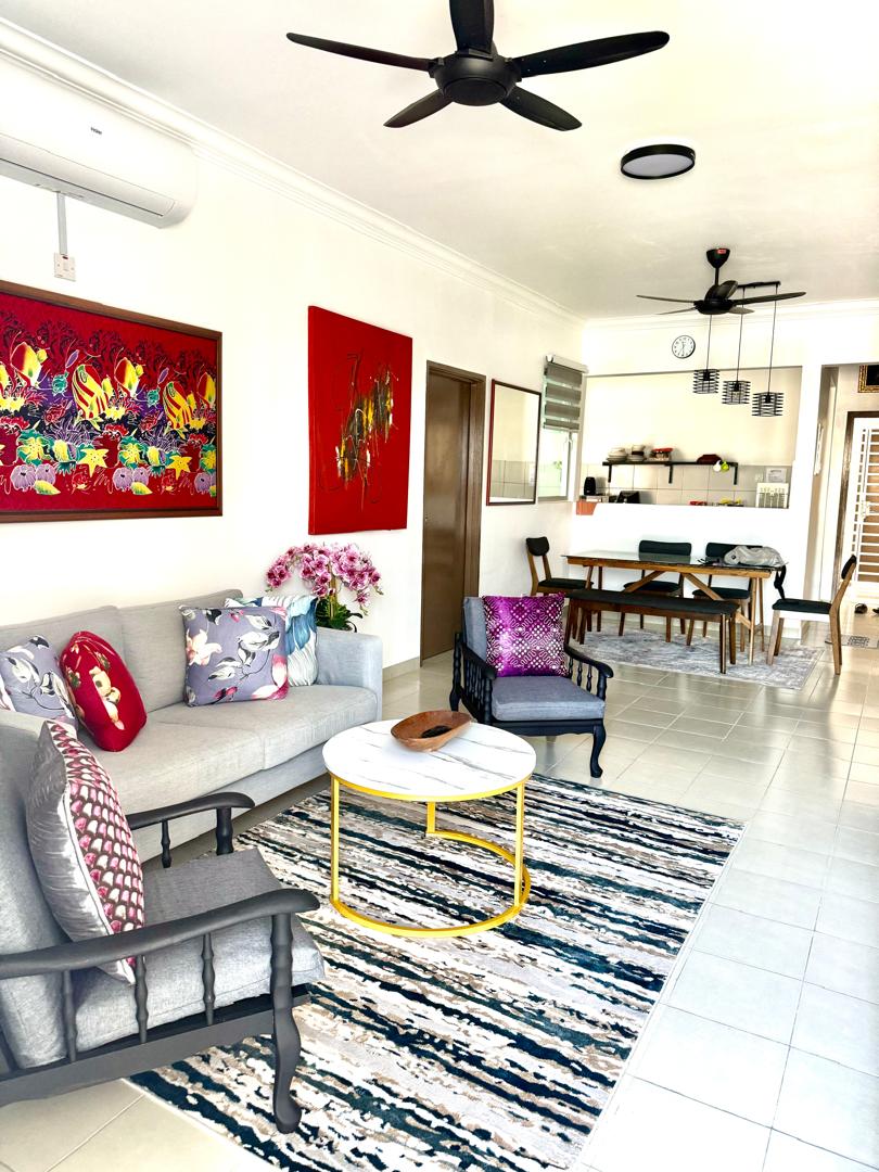 A living area features a light-colored sofa with decorative pillows and two black armchairs. A round, gold-accented coffee table sits on a patterned rug. Colorful artworks adorn the walls, and a dining area with a wooden table and chairs is visible in the background.